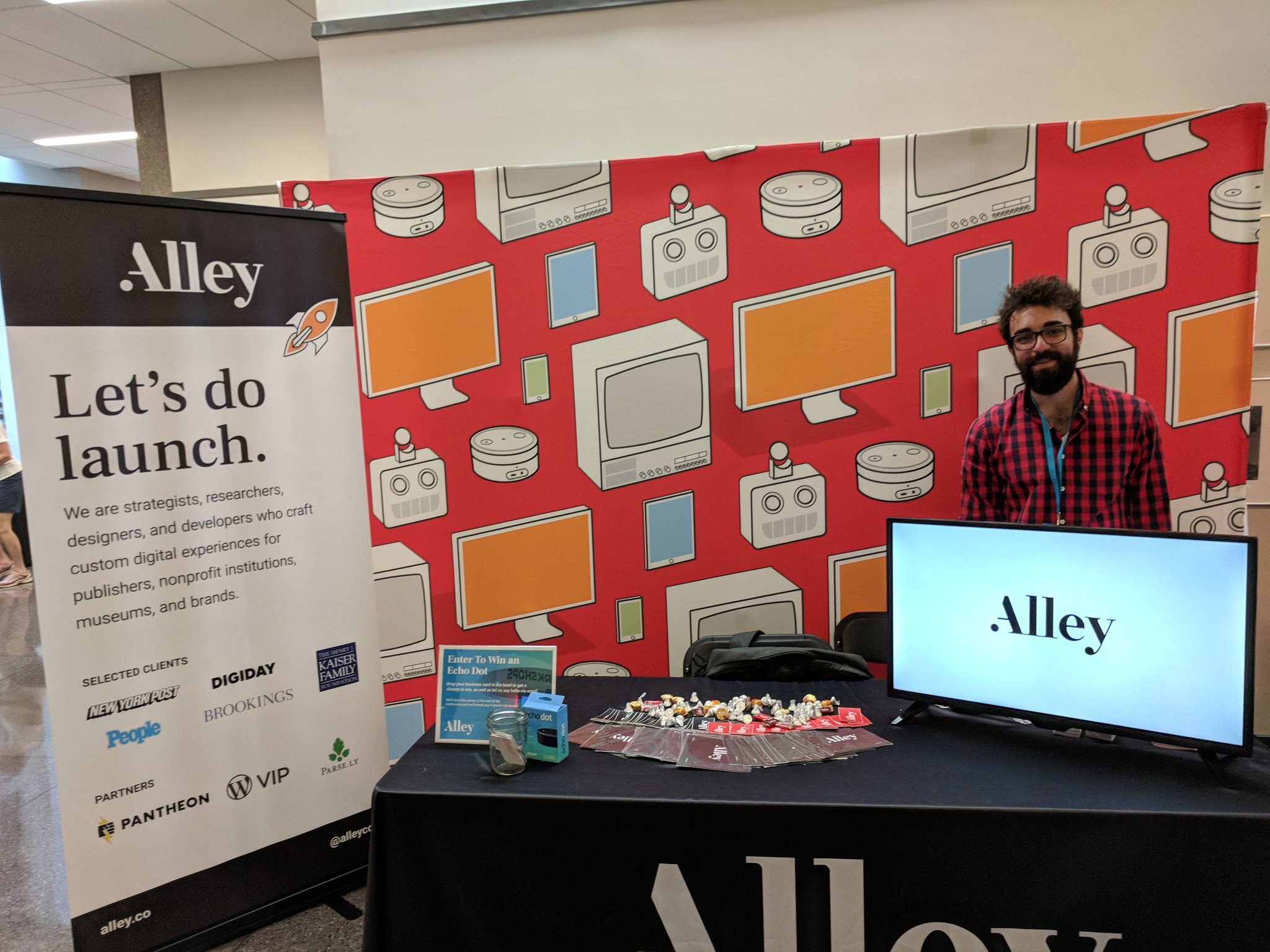 The Alley conference booth with a red  background with computers and tech devices, a television screen with the Alley logo, and a banner, as well as a White man in a plaid shirt smiling at the camera