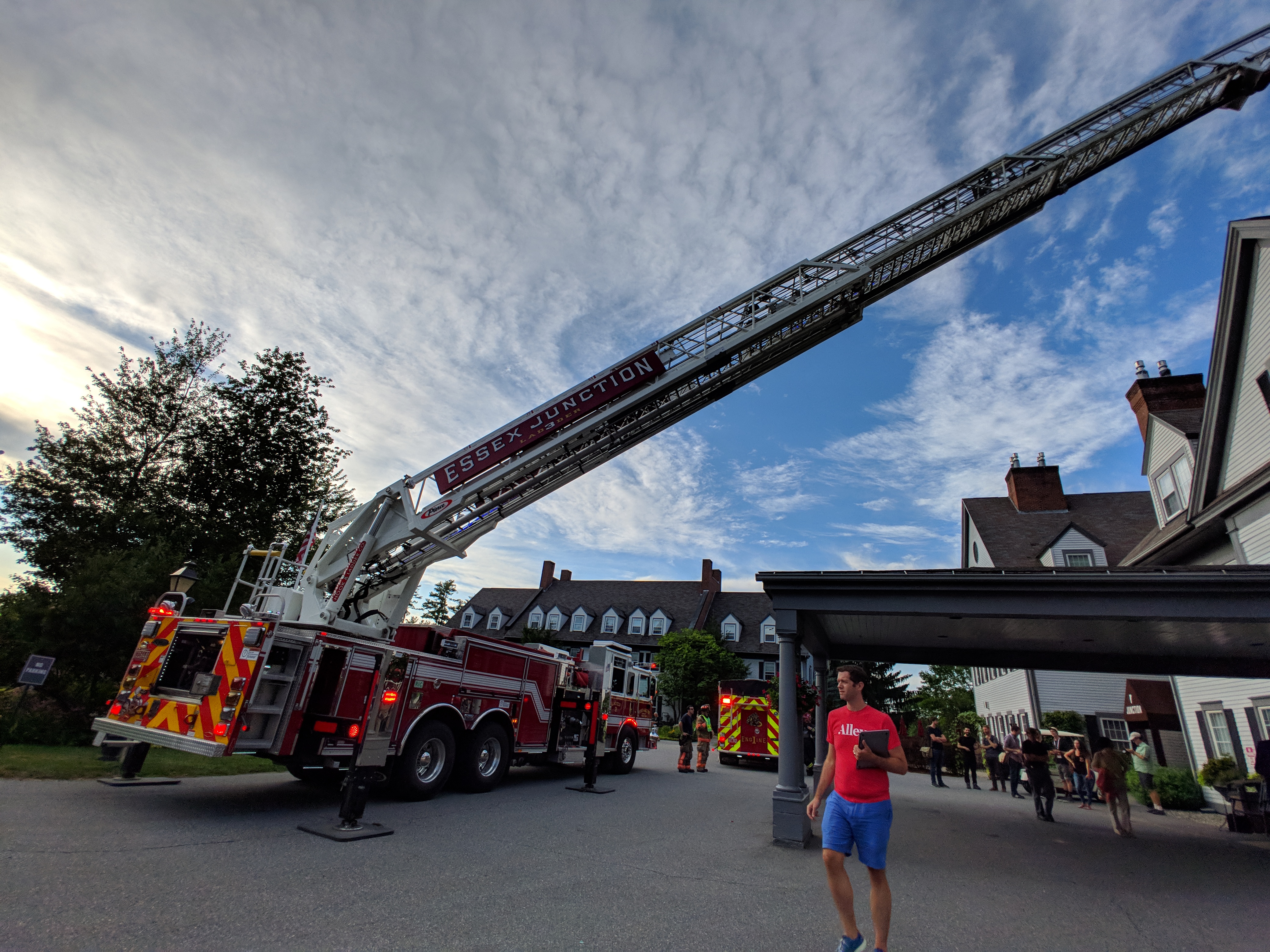 A fire truck with a long ladder fully extended to reach the top of the building next door while a White man with a red Alley shirt and blue shorts walks in front