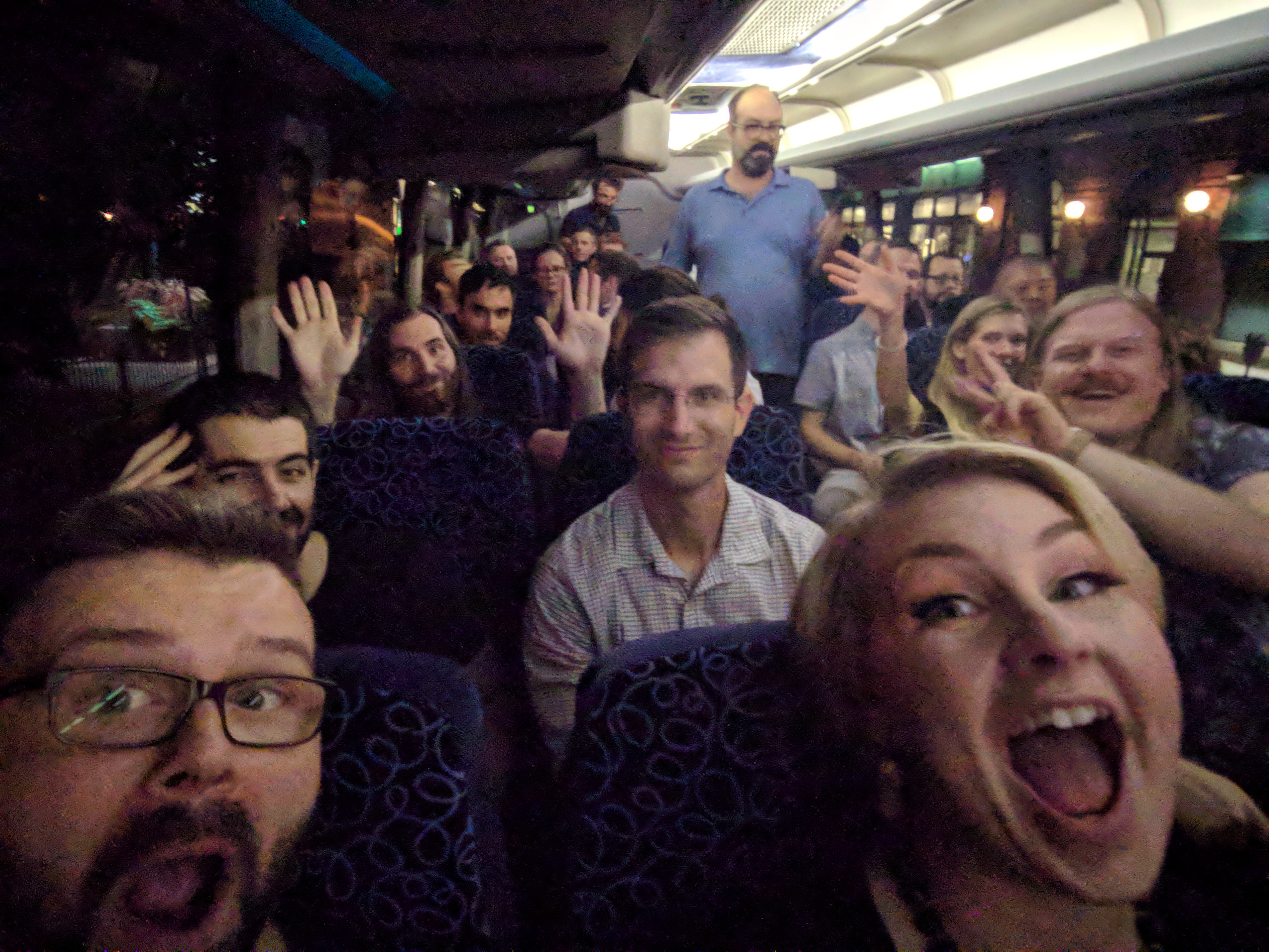 A selfie of many team members on a bus smiling and waving their hands