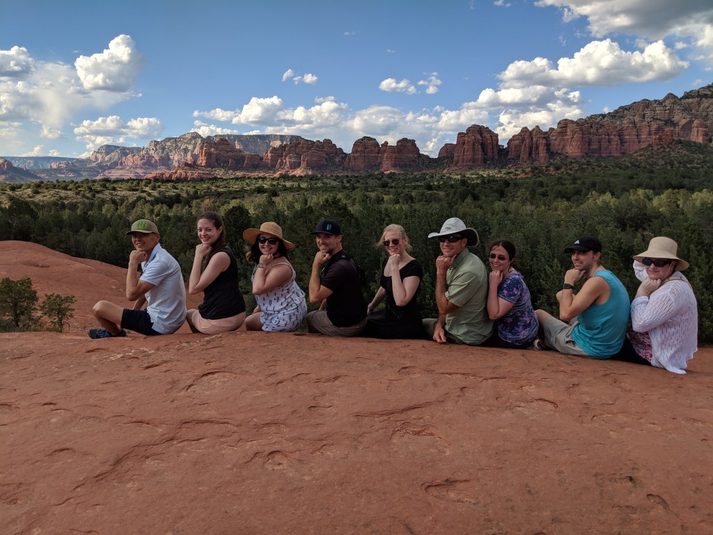 Team VIP sitting in a row all holding the same pose with their fists under their chins as though pondering something. Backdrop against the red rocks at Sedona