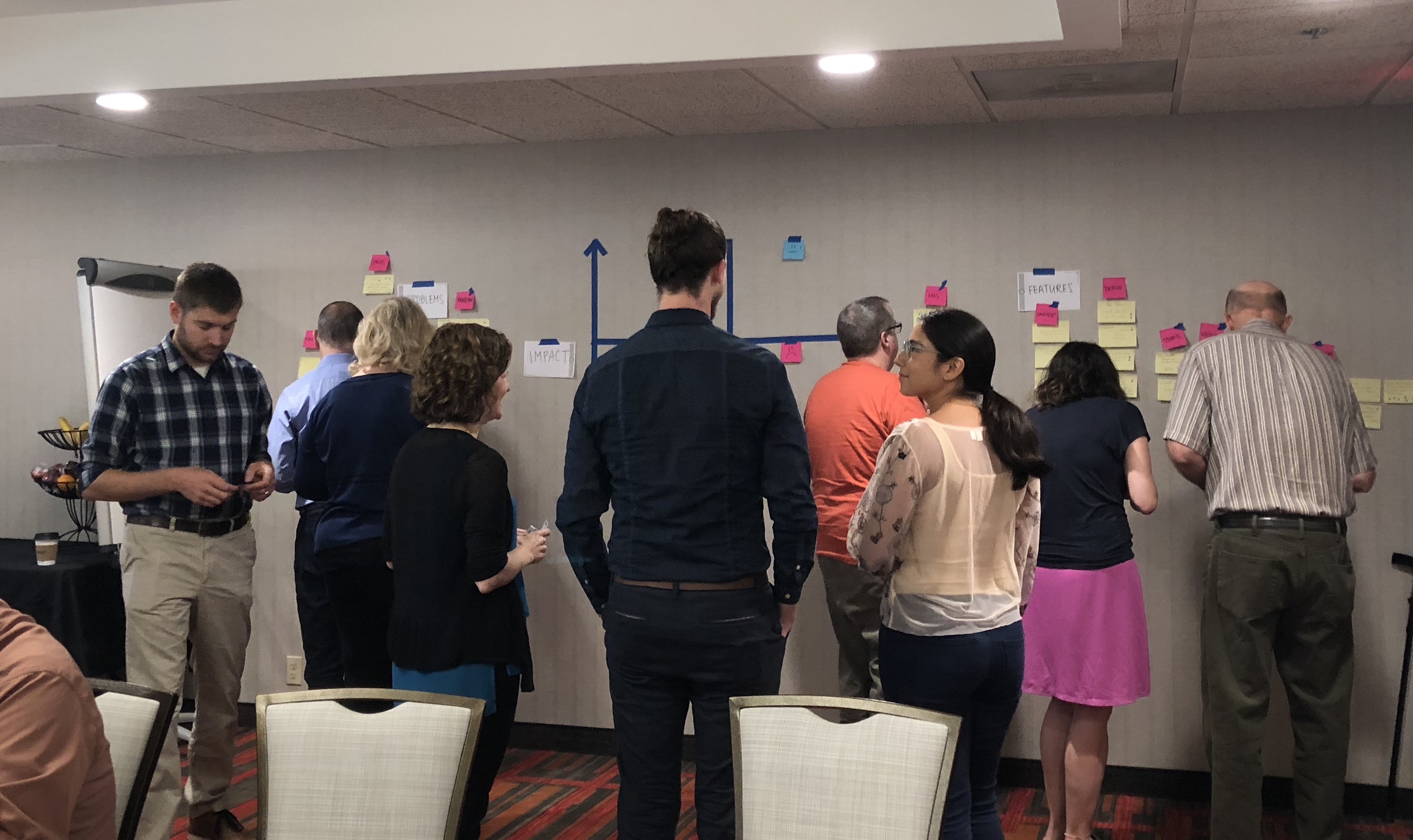 A large group of people looking at a display of post its and tape on the wall of a conference room