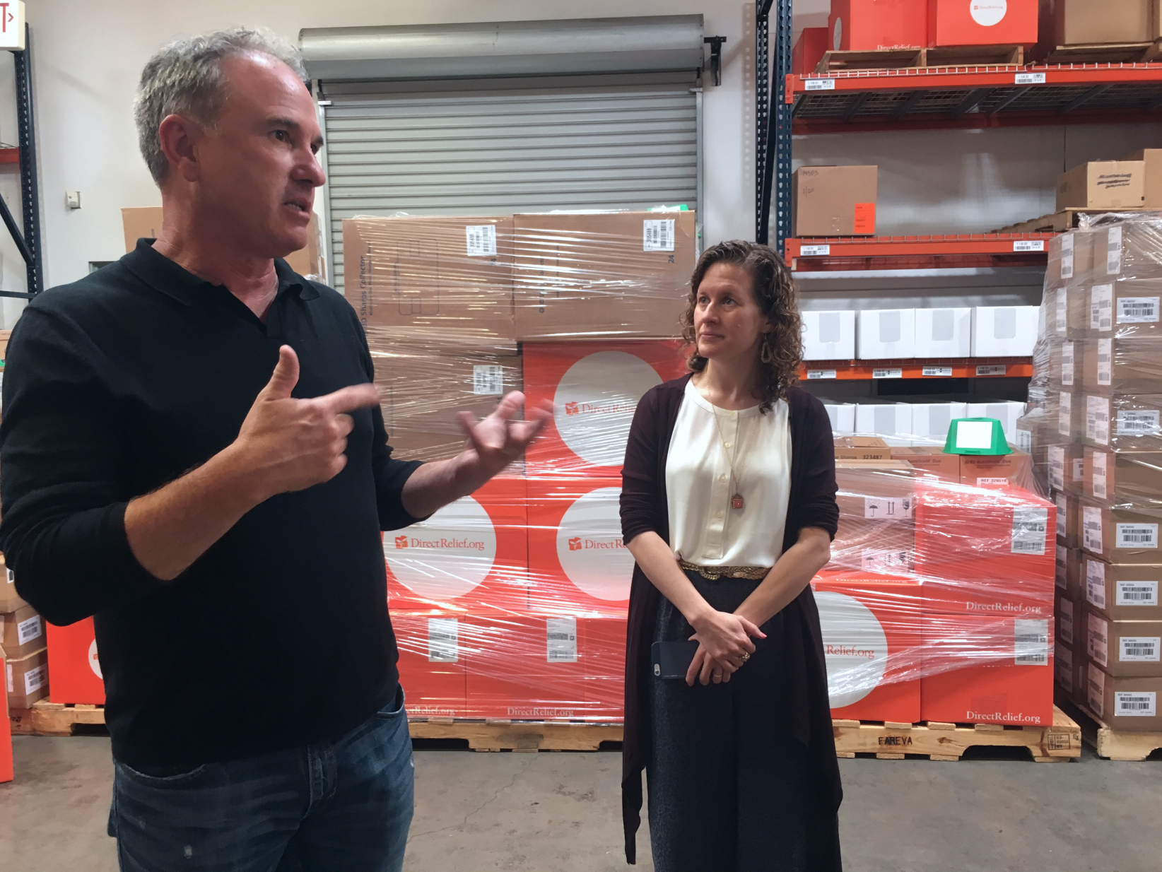 A woman listening to a gesturing man in a large warehouse in front of many shrinkwrapped orange boxes with the Direct Relief logo on them