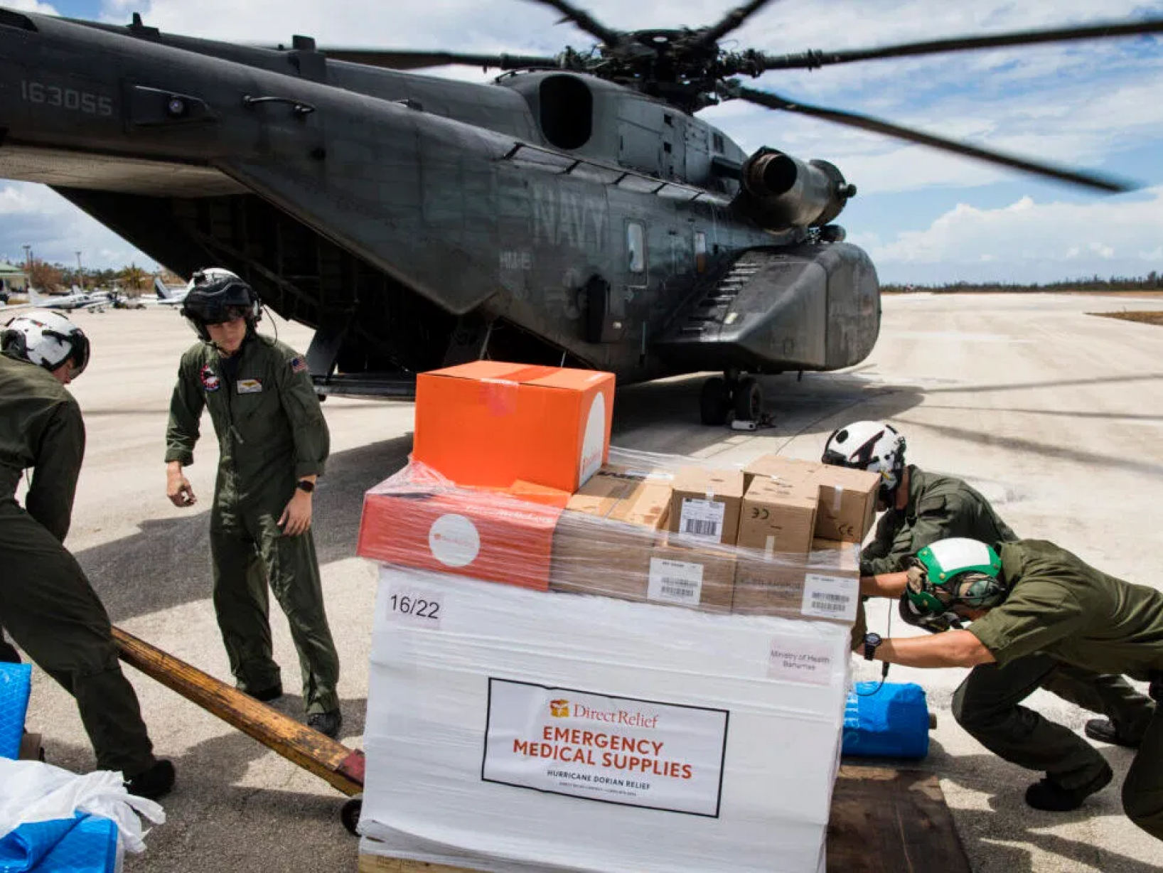 Four men in helmets pushing a pallet labelled emergency medical supplies into a large black Navy helicopter