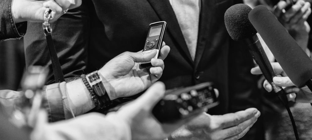 A black and white photograph that is zoomed in on the hands of reporters holding recording devices.