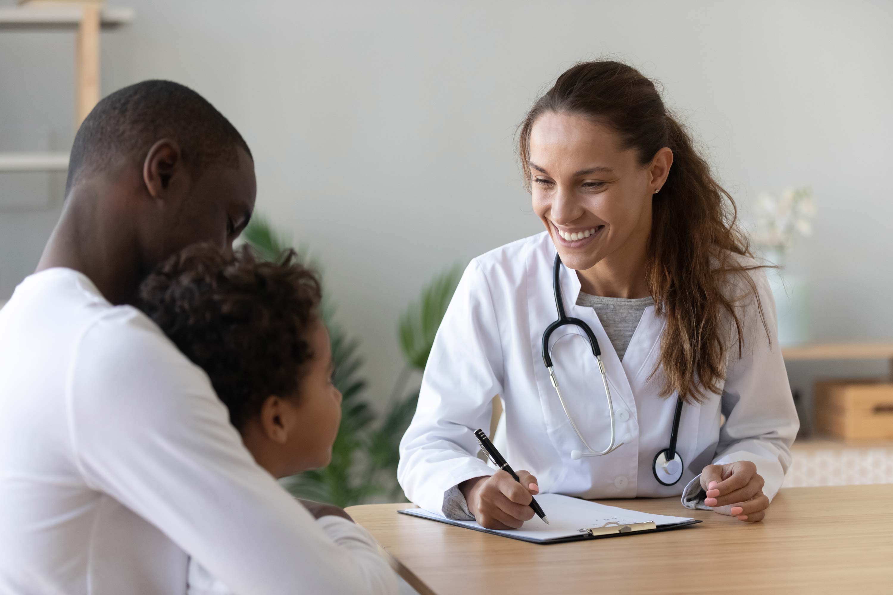 Image of a woman doctor smiling at a child patient and his father. The child is sitting on the father's lap, and they are facing the doctor who is seated at her table.