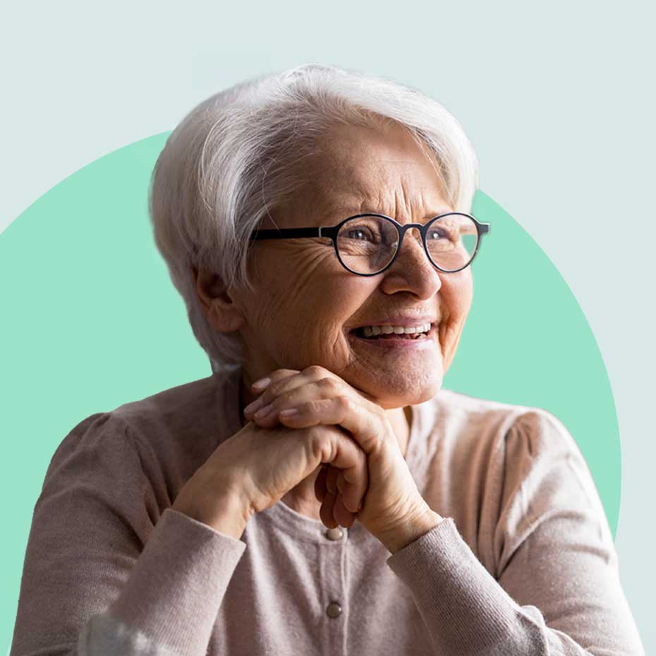 An older woman with short white hair and glasses smiles warmly, resting her hands under her chin against a soft green background.